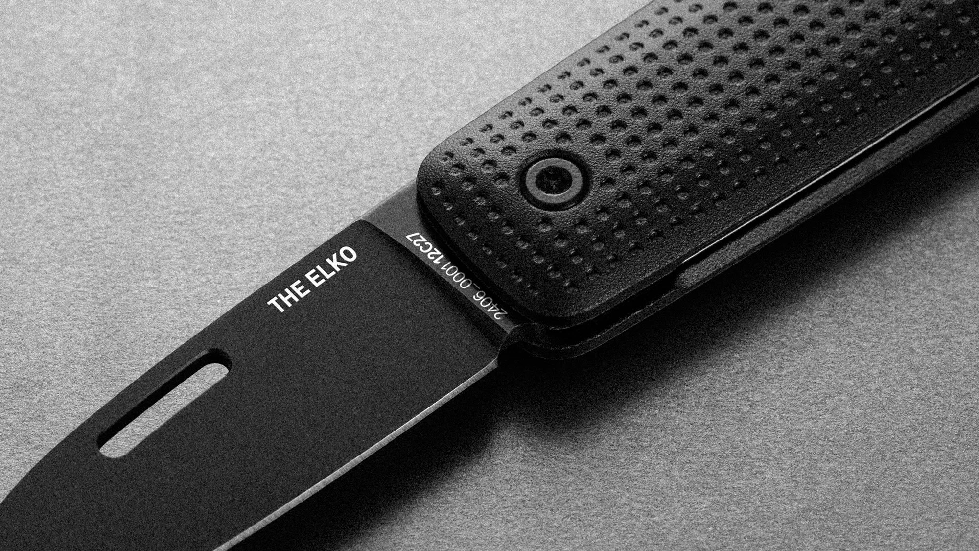 Black folding knife blade labeled "THE ELKO" with textured black handle on gray surface.