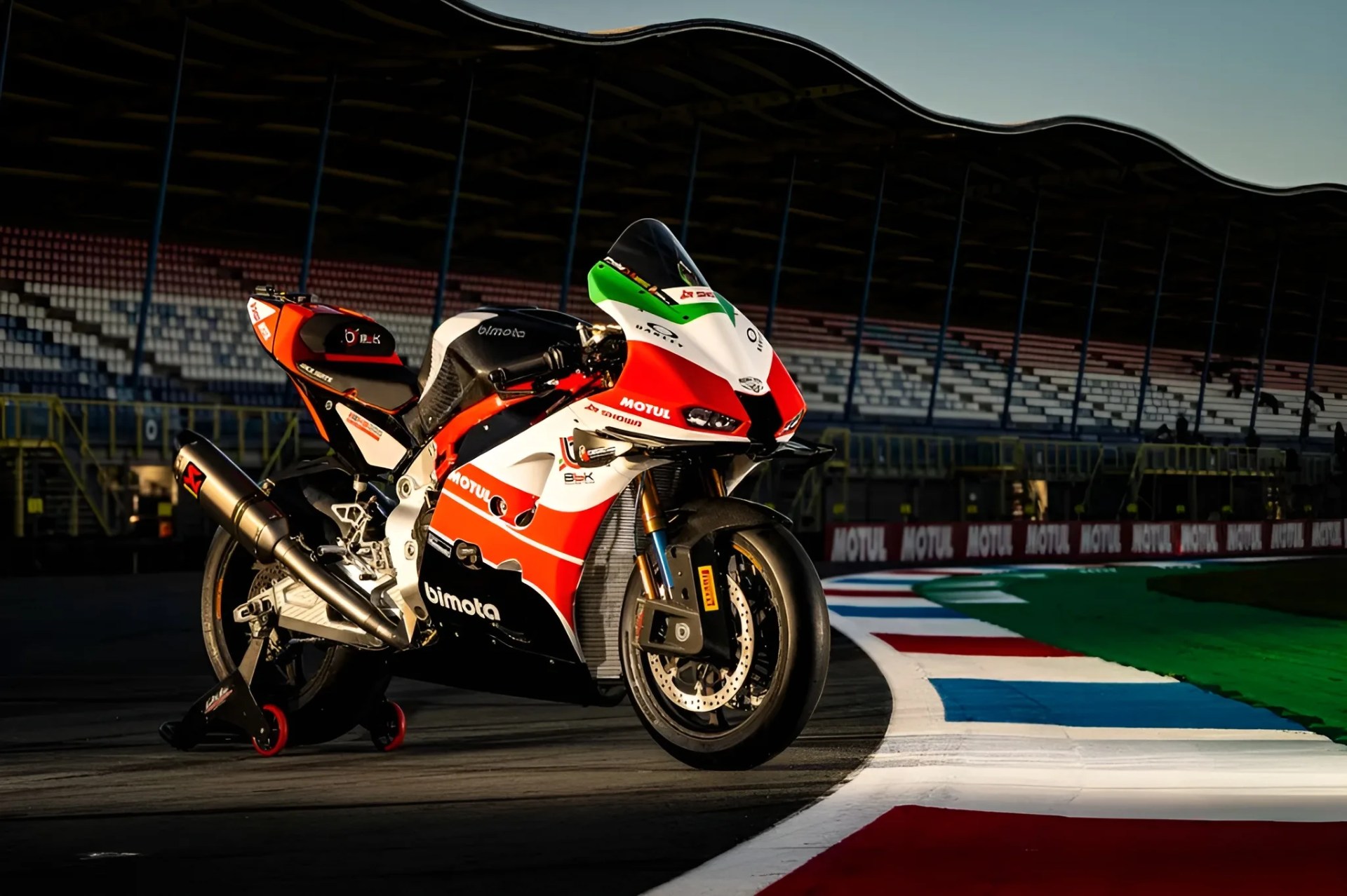 Red, white, and black Bimota racing motorcycle on a racetrack with grandstands in the background.