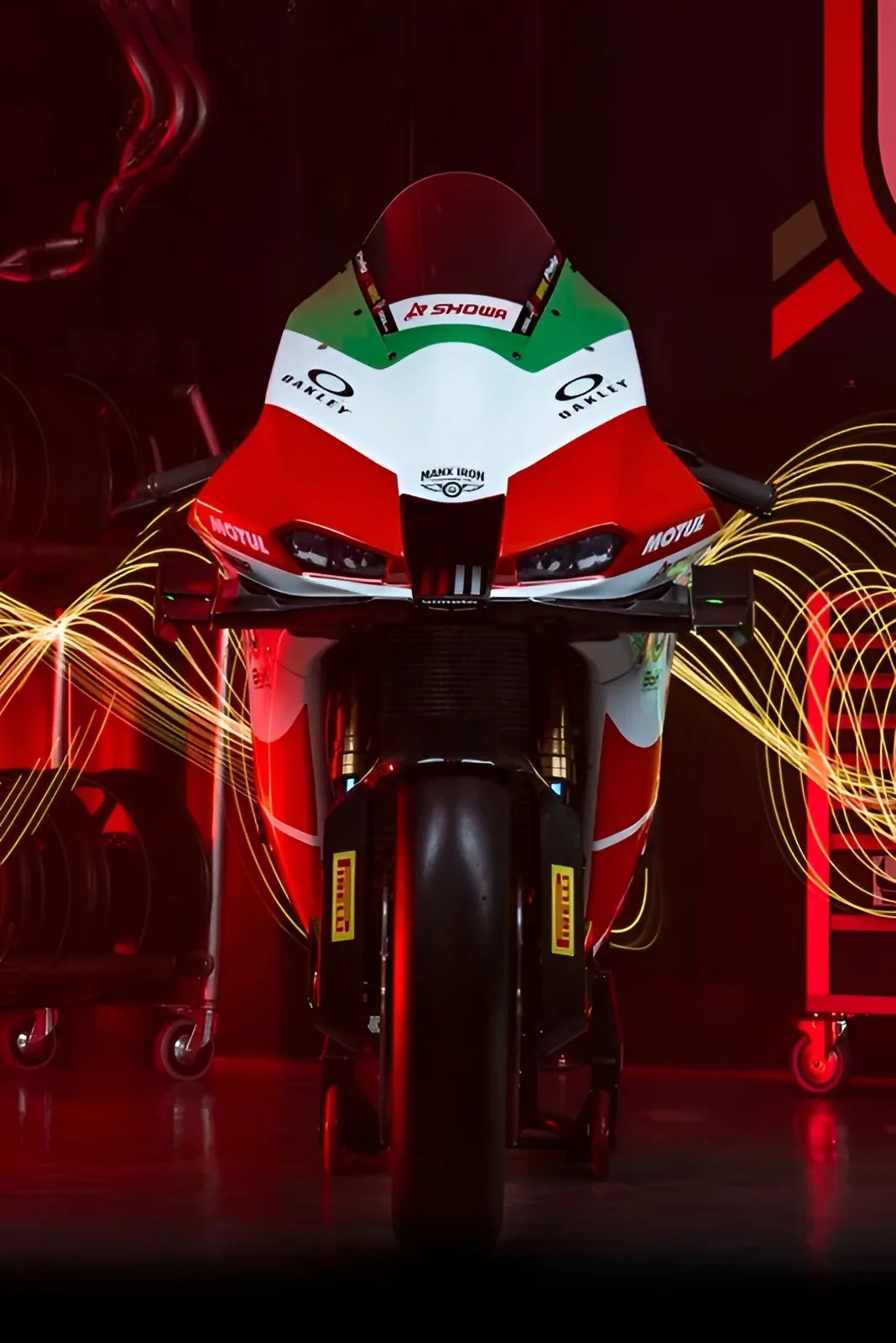 Front view of a red, white, and green racing motorcycle with sponsor logos in a dark room with red lighting.