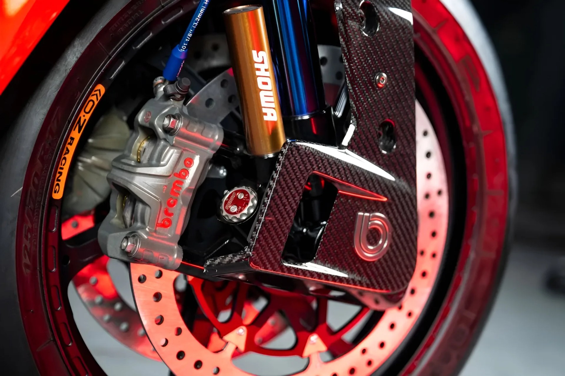 Close-up of a motorcycle front wheel with Brembo brake caliper, Showa suspension, carbon fiber fork guard, and OZ Racing tire.