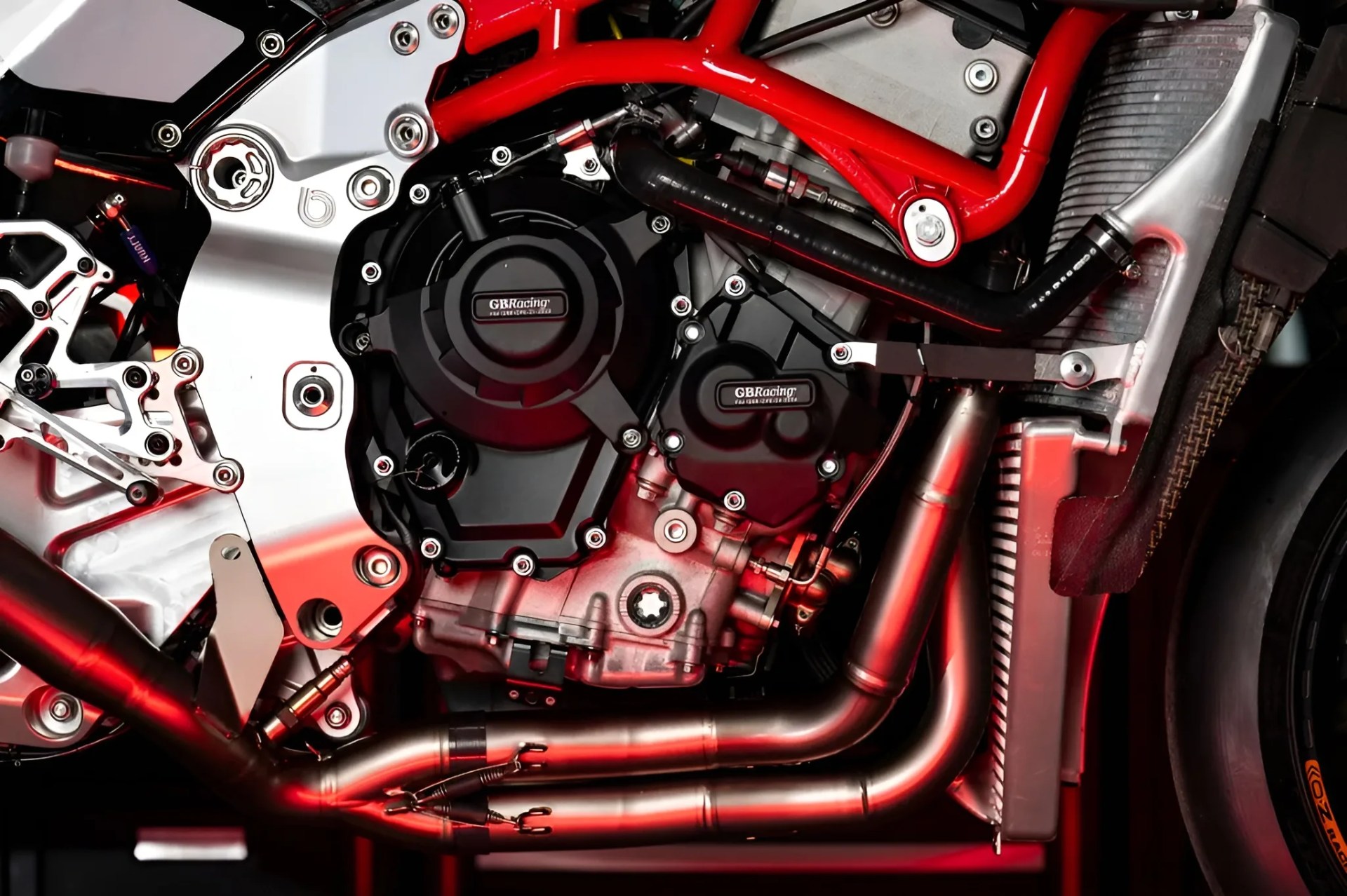 Close-up of a motorcycle engine with black GB Racing covers, red frame, and silver exhaust pipes.
