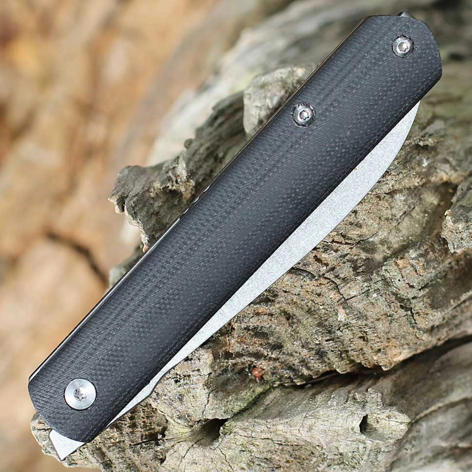 Black folding knife with textured handle resting on rough tree bark.