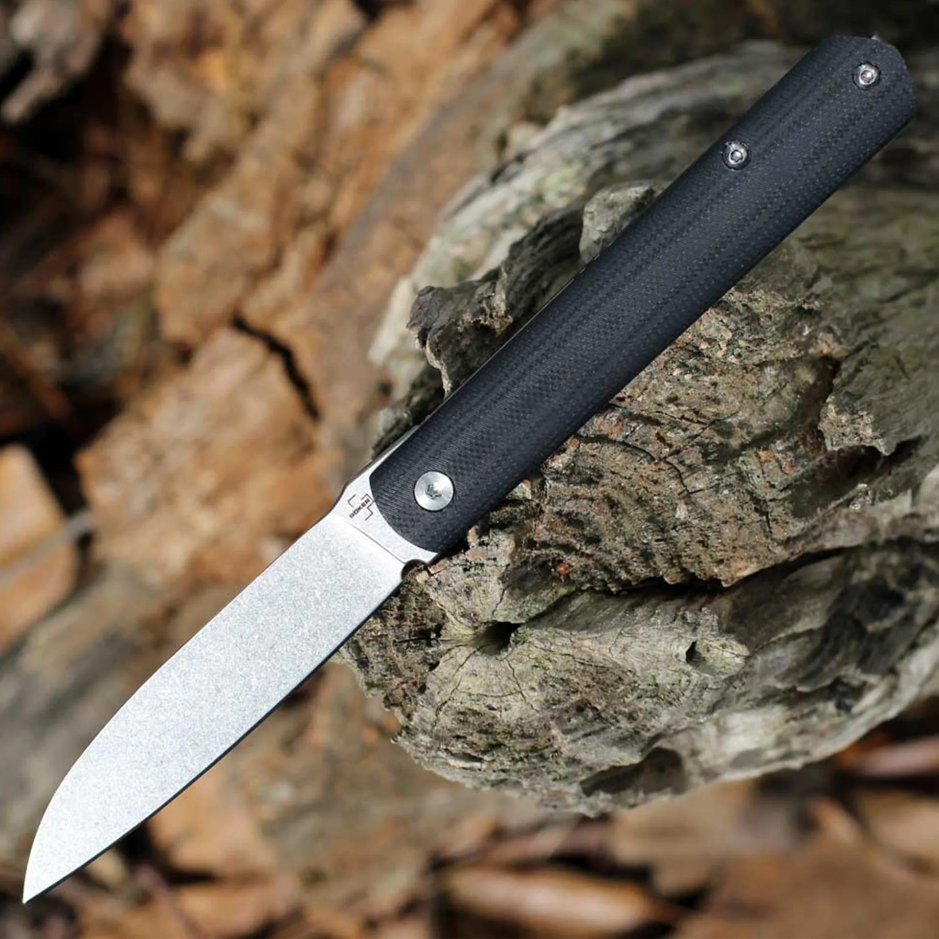 Folding knife with a stonewashed blade and black textured handle resting on a tree stump.