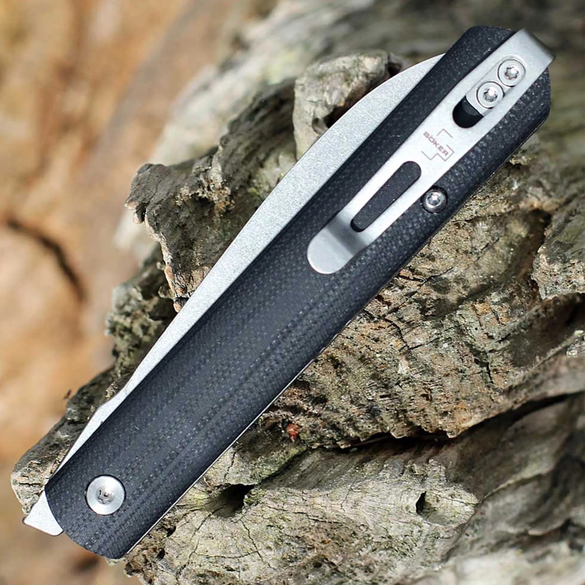 Folded black and silver Boker pocket knife resting on rough tree bark.
