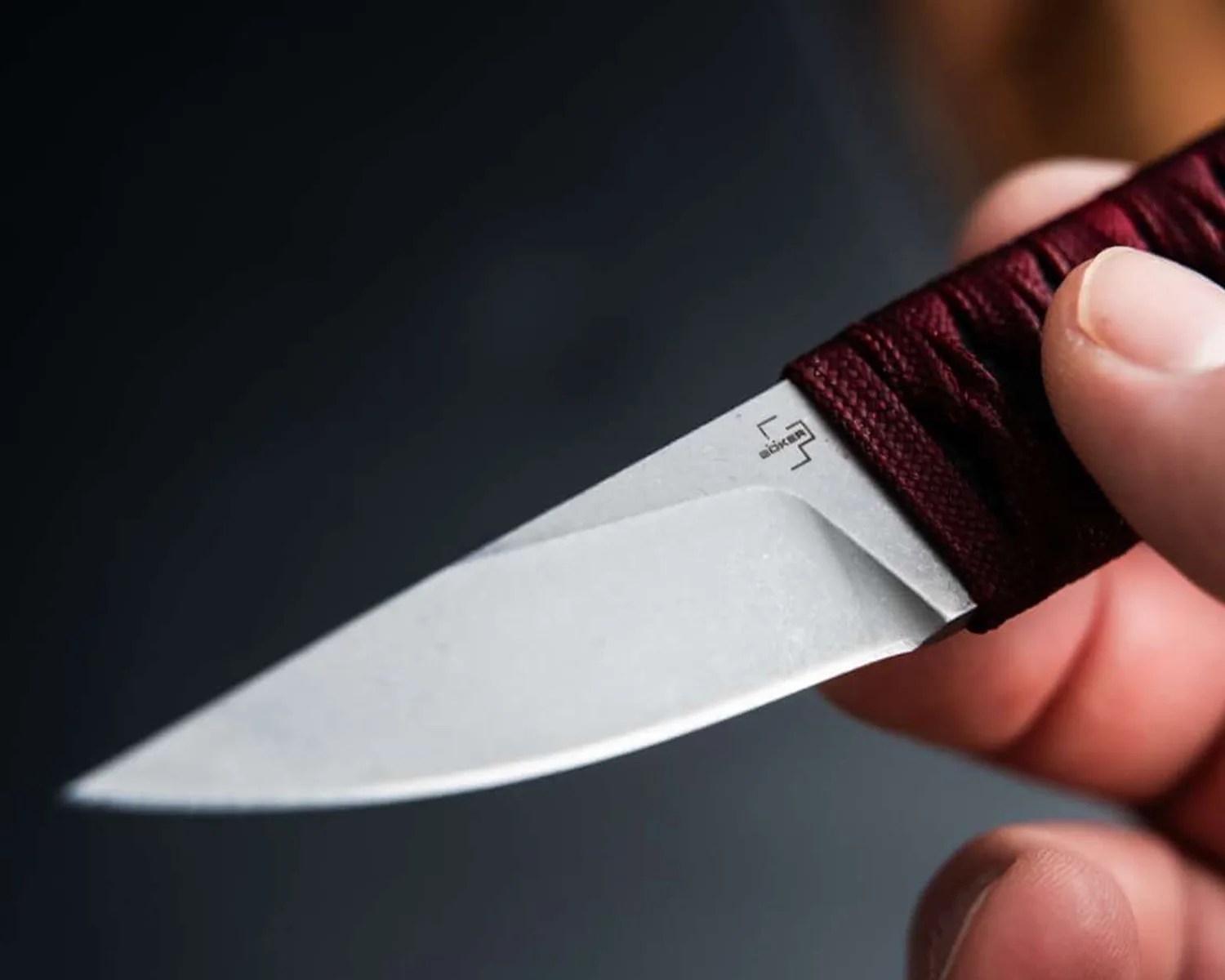 Close-up of a small fixed-blade knife with a stonewashed steel blade and a dark red wrapped handle held in a hand.