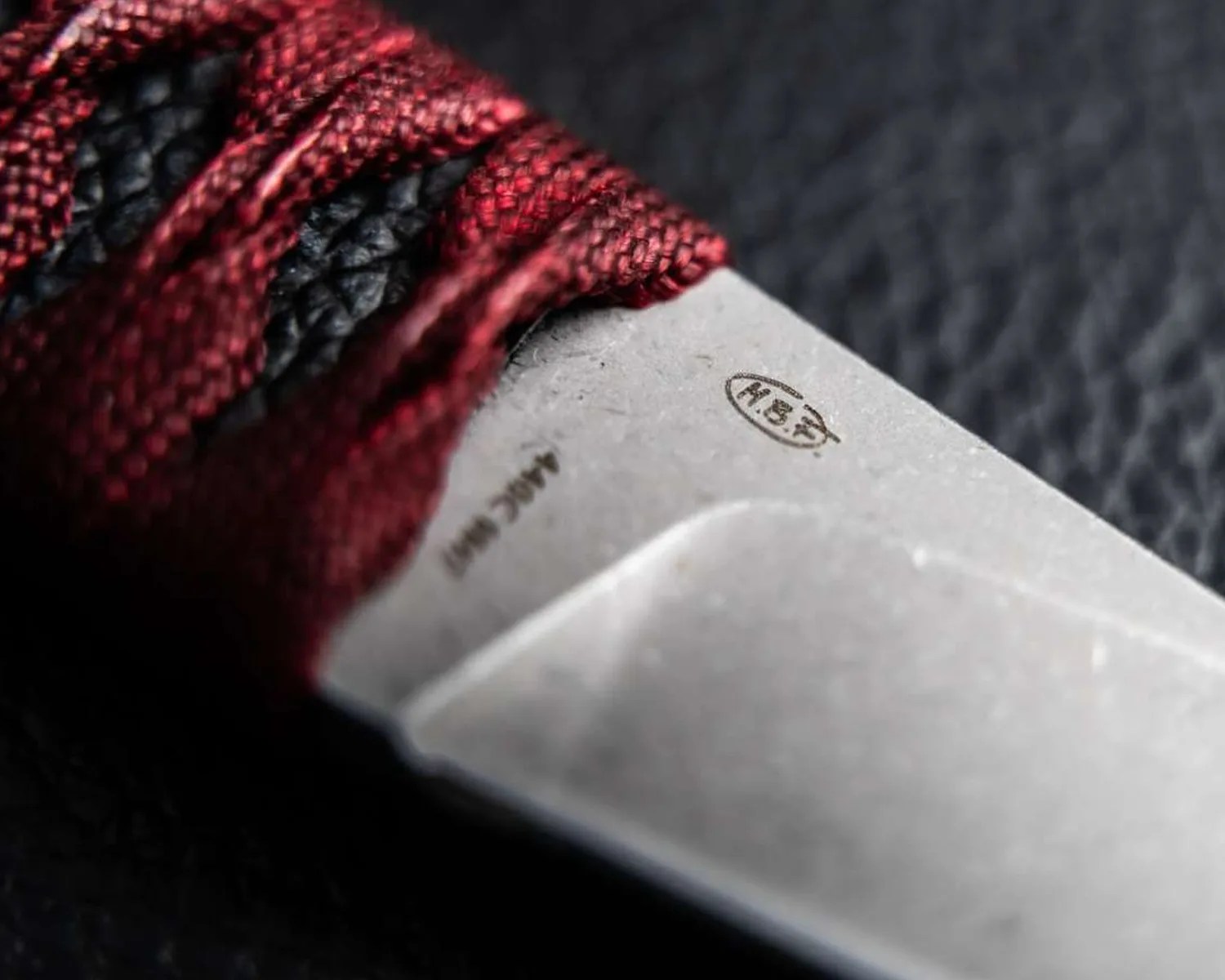 Close-up of a knife blade with red and black cord-wrapped handle and "M.B.F" engraved on the blade.