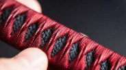 Close-up of a hand holding a red and black textured handle wrapped in a traditional Japanese tsuka-ito style.