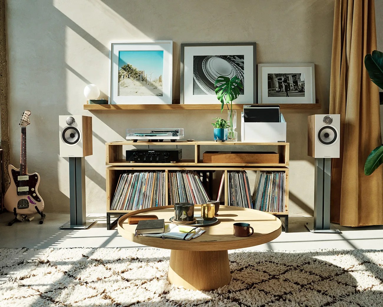 Wooden media console with vinyl records, black amplifier, and turntable, flanked by white speakers on stands in sunlit room.