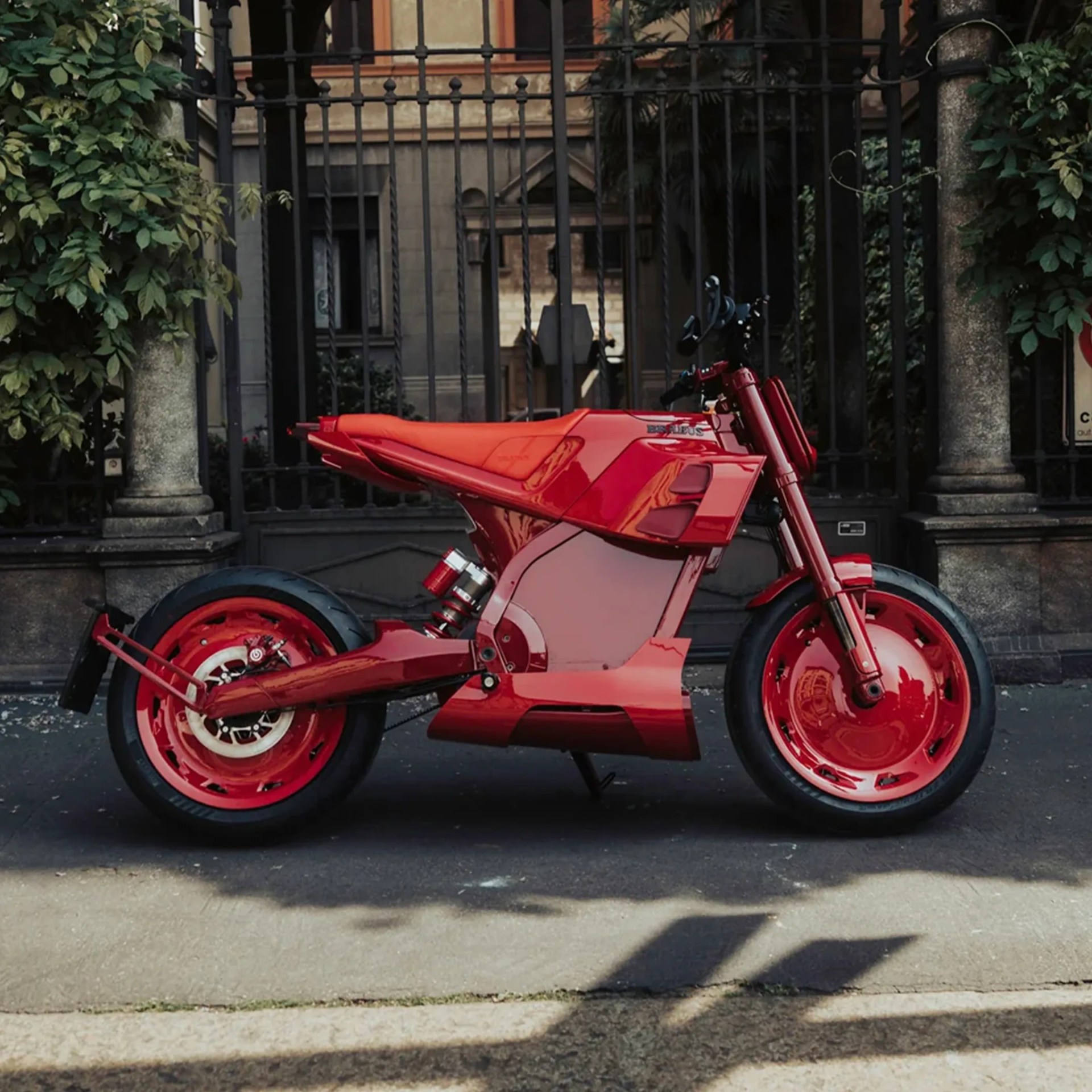 Red futuristic electric motorcycle parked on a street with a black iron fence and greenery in the background.