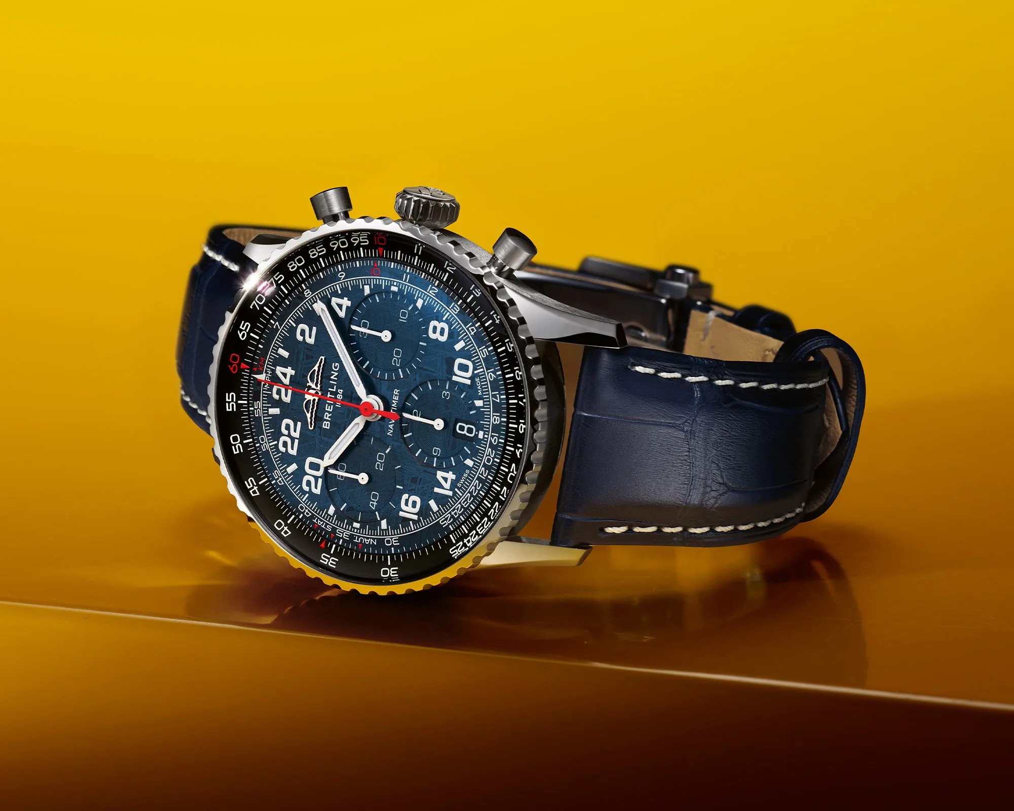 Breitling Navitimer wristwatch with blue dial, chronograph subdials, and blue leather strap on yellow surface.