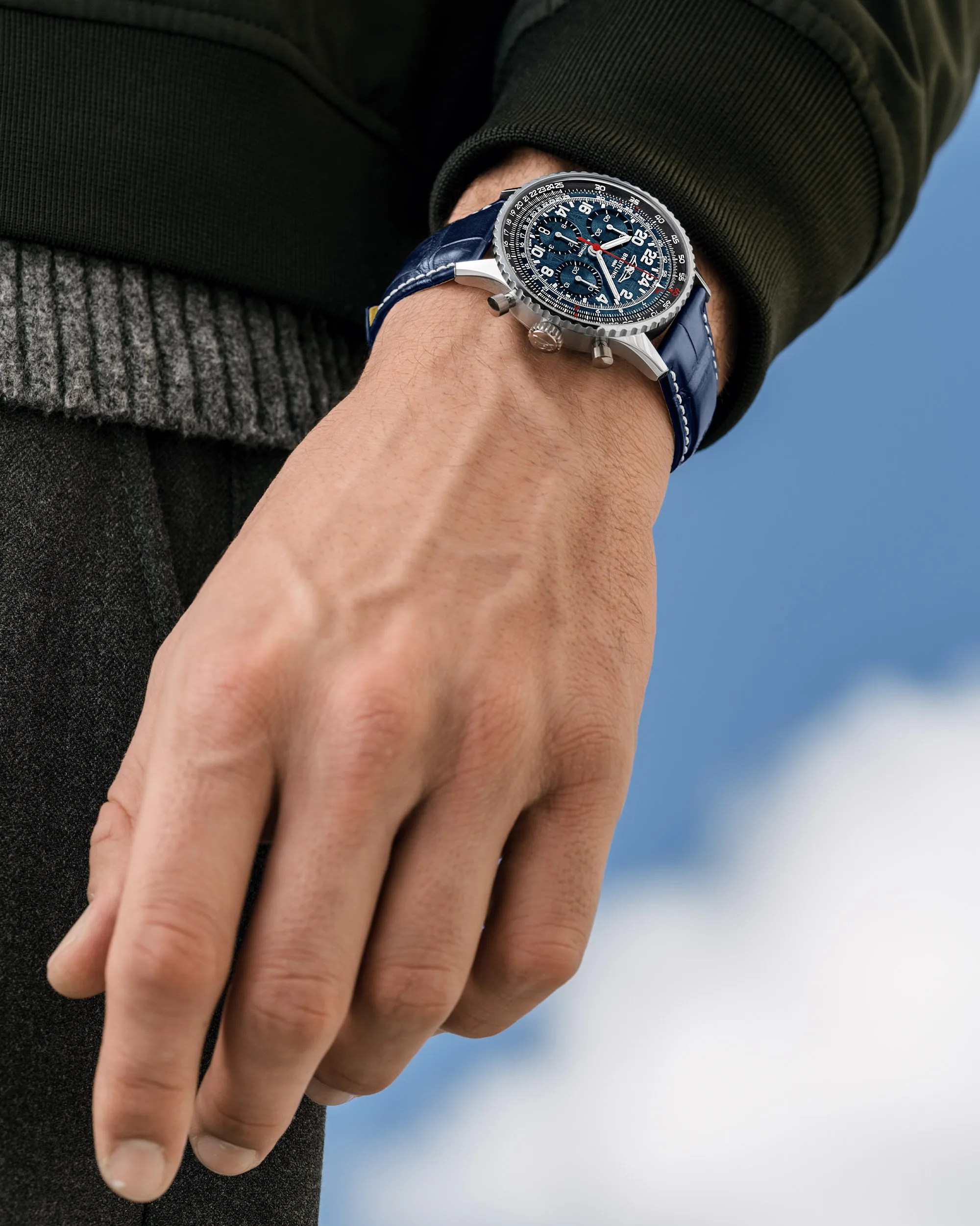 Silver chronograph wristwatch with a blue dial and blue leather strap worn on a wrist against a blue sky background.
