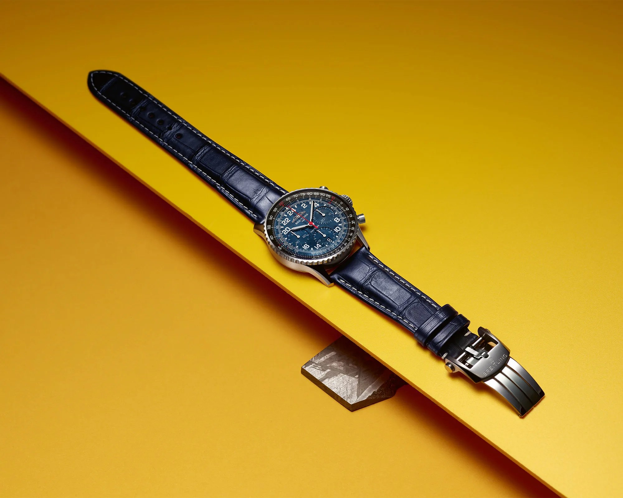 Blue leather-strap wristwatch with a detailed blue dial and silver clasp on a yellow surface.