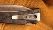 Close-up of a folding knife handle with a camo pattern and a metal pocket clip attached by two screws.