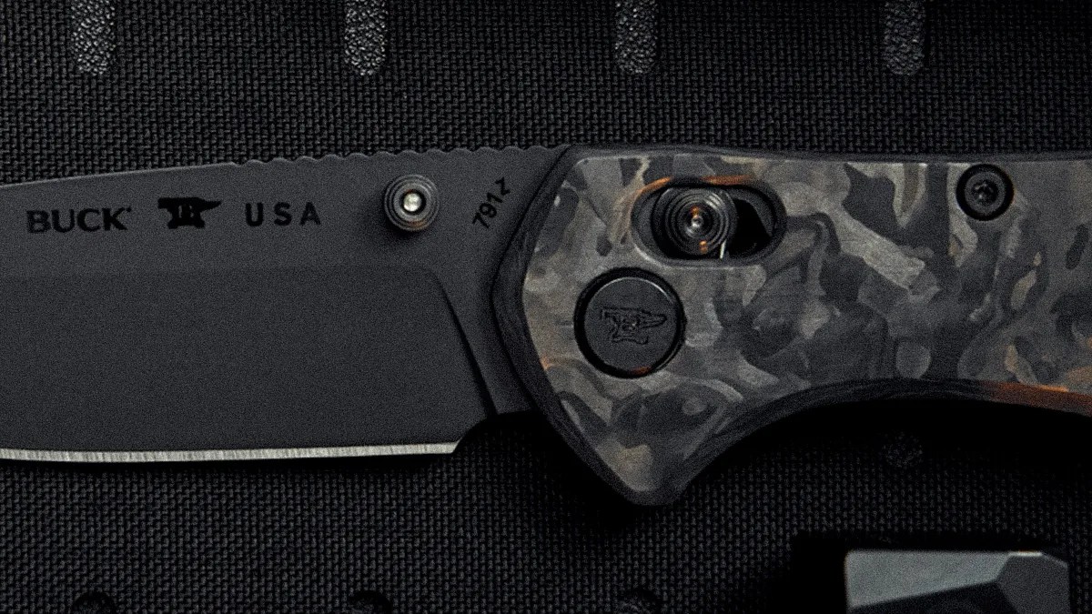 Black folding knife blade with "BUCK USA" text and a camo-patterned handle on a textured black surface.