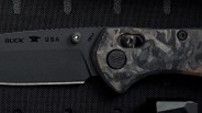 Black folding knife blade with "BUCK USA" text and a camo-patterned handle on a textured black surface.