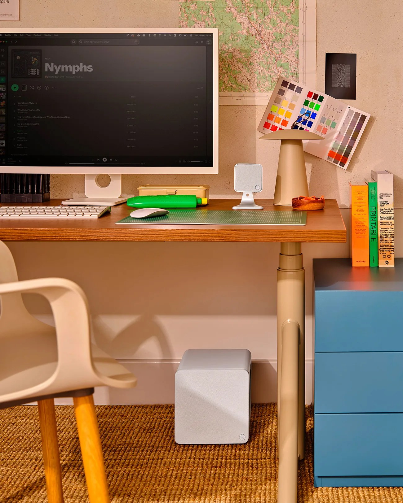 Home office desk setup with a large monitor displaying a music playlist, beige chair, green cutting mat, white speaker, and colorful books on a blue drawer unit.