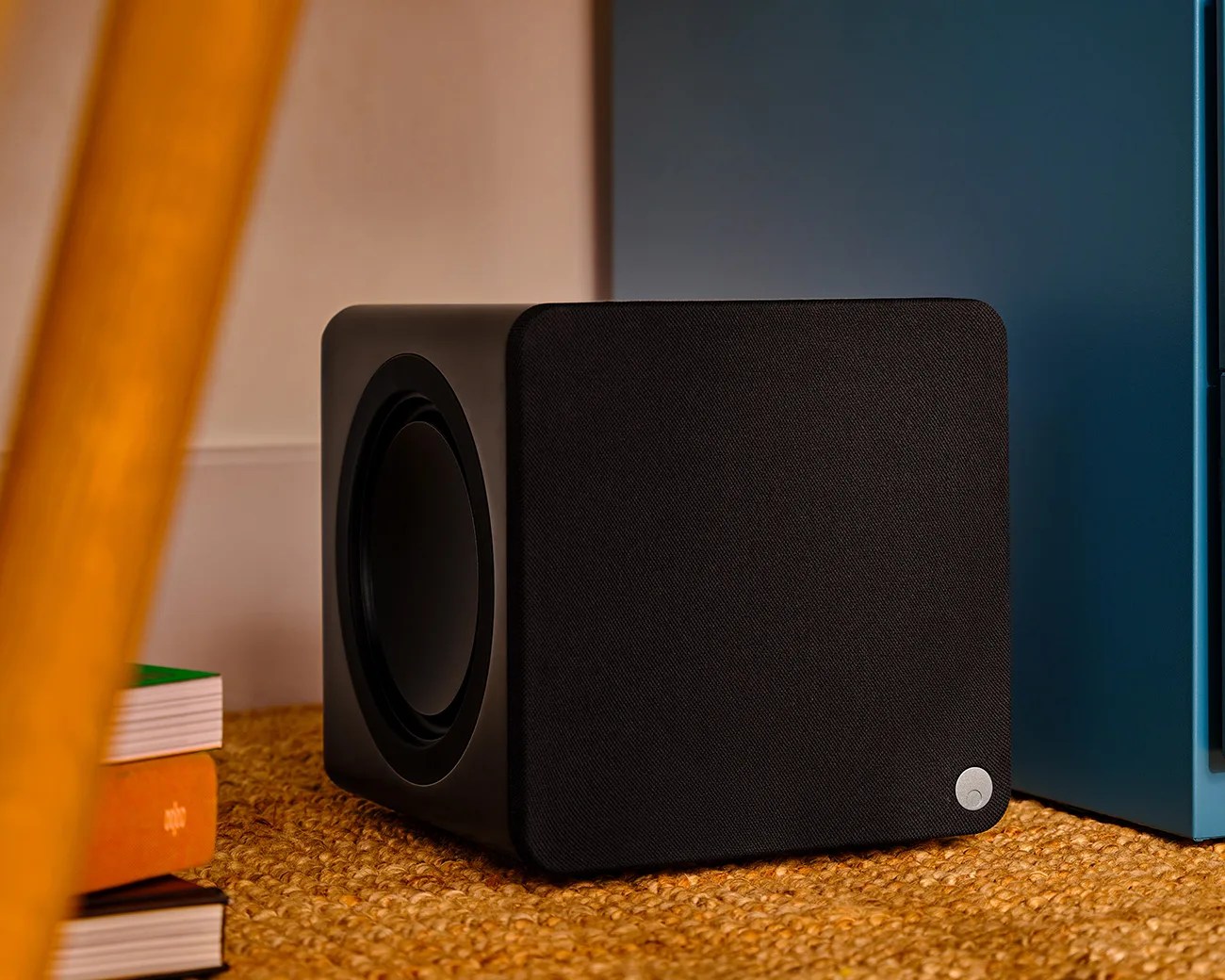 Black cube-shaped speaker with fabric front and side driver on a woven surface next to books and a blue cabinet.