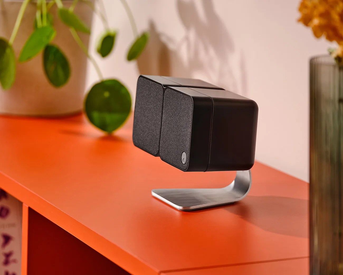 Pair of black rectangular speakers with fabric fronts on a curved silver metal stand on an orange surface.