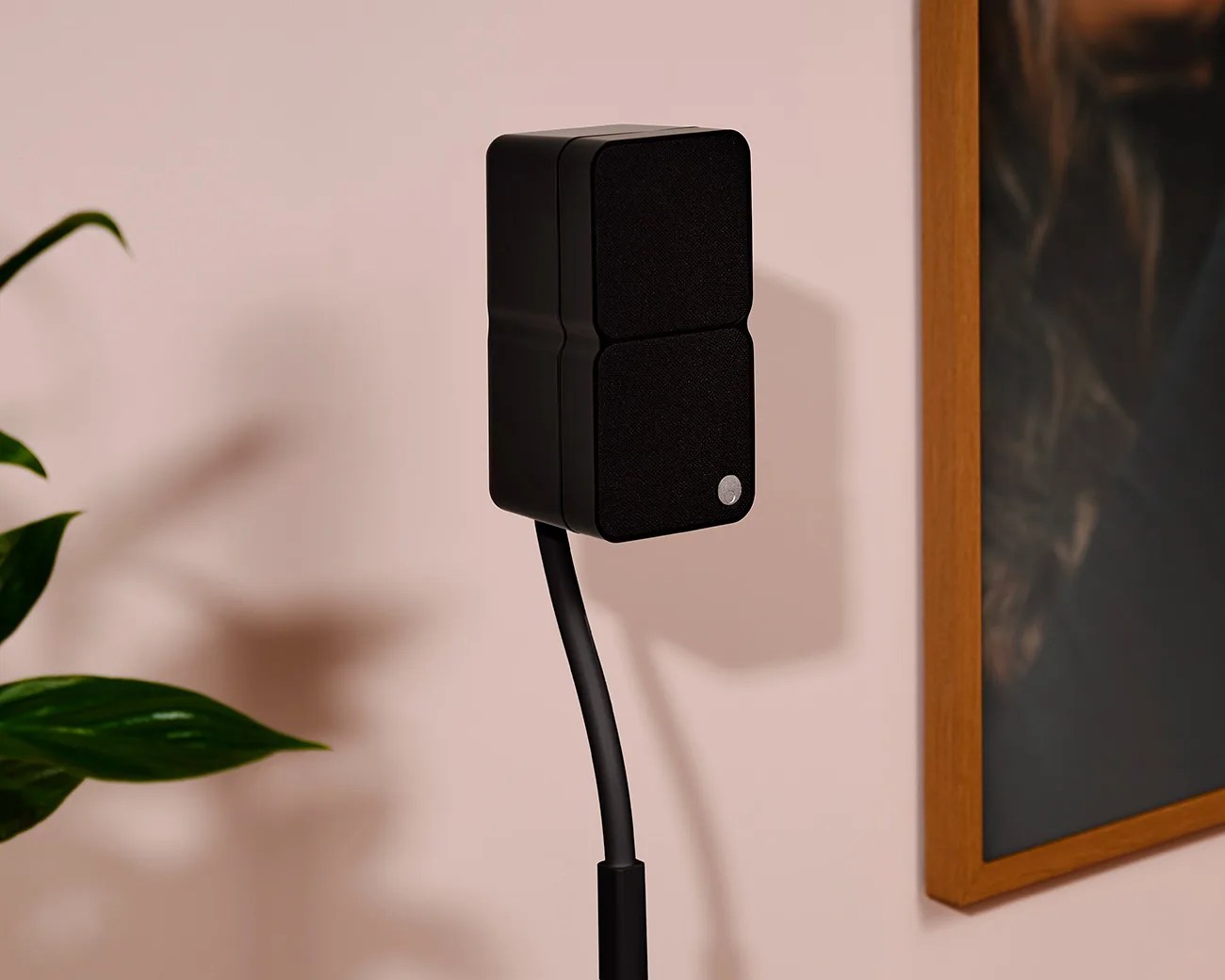 Black rectangular speaker with fabric front mounted on a flexible black stand against a beige wall.