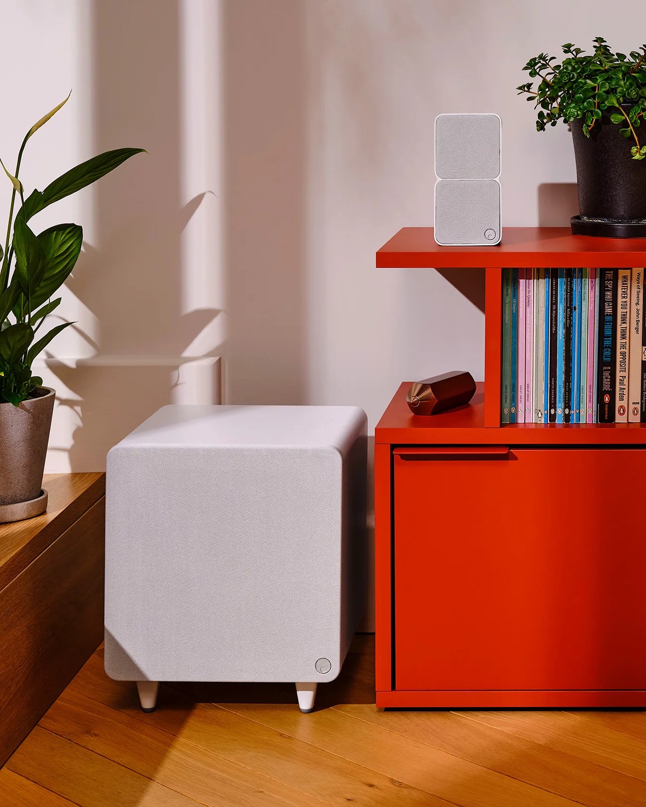 White fabric-covered subwoofer on wooden floor next to a red bookshelf with a small white speaker and books on top.