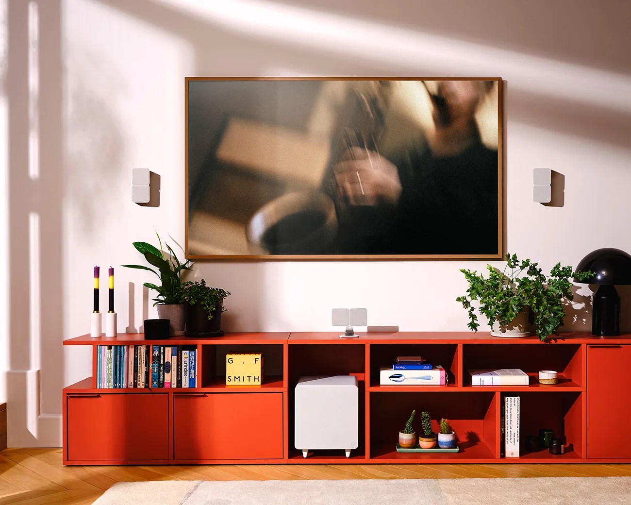 Red modular shelving unit with books, plants, candles, and a black dome lamp under a framed blurry photo on a white wall.