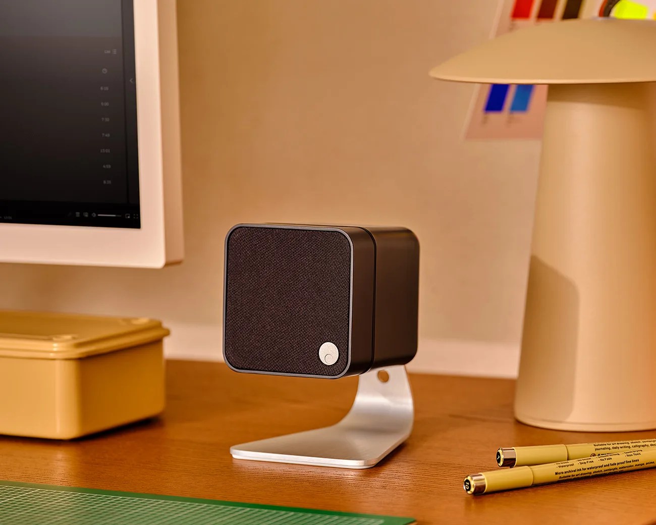 Black cube speaker with fabric front and silver stand on a wooden desk near a computer monitor and beige lamp.