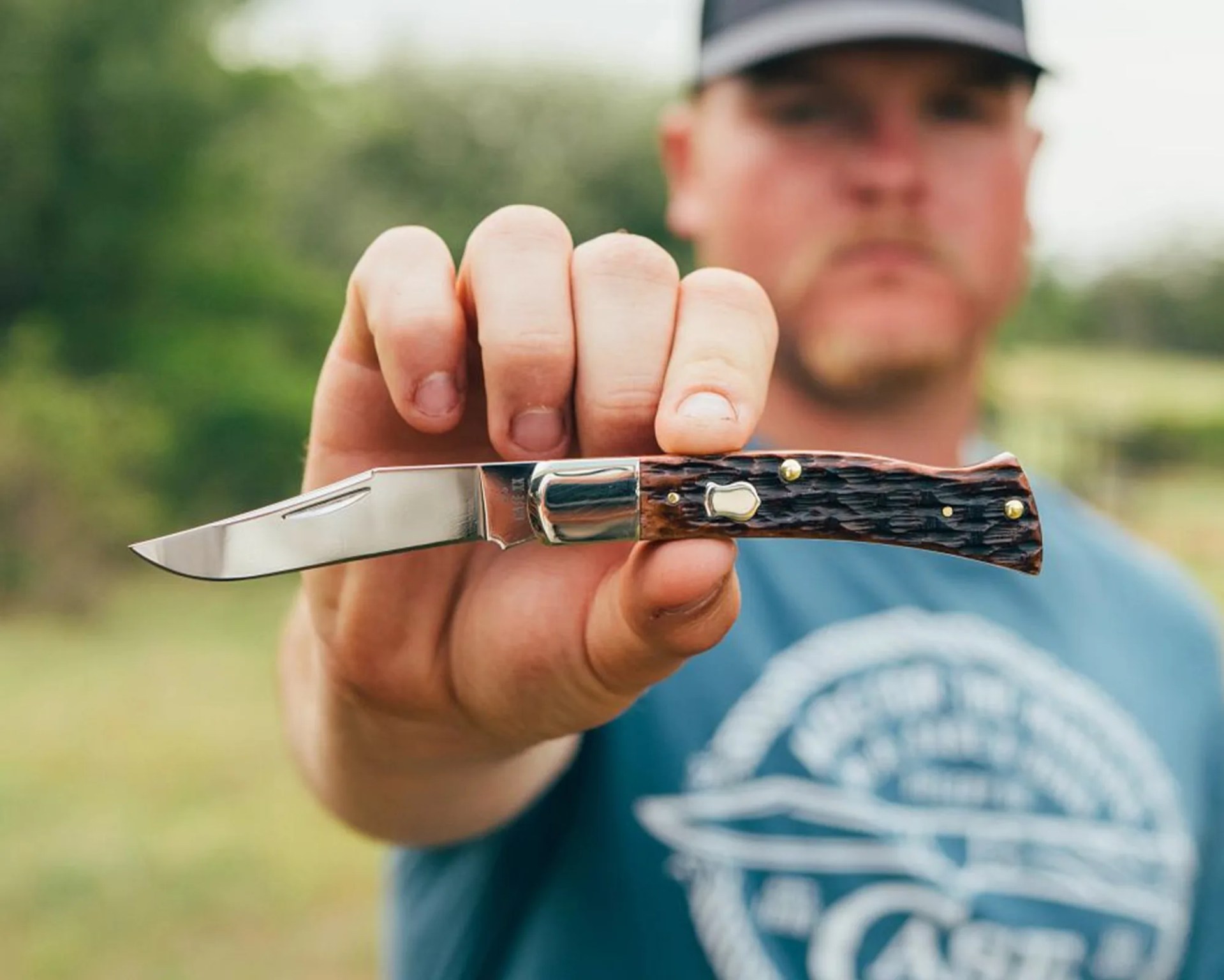 Hand holding a folding pocket knife with a polished blade and textured dark wooden handle outdoors.