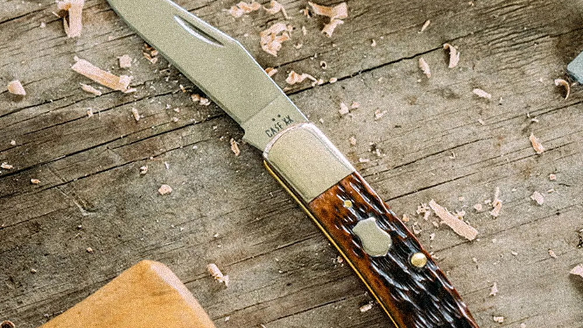 Folding pocket knife with a polished metal blade and a textured brown handle on a wooden surface with wood shavings.