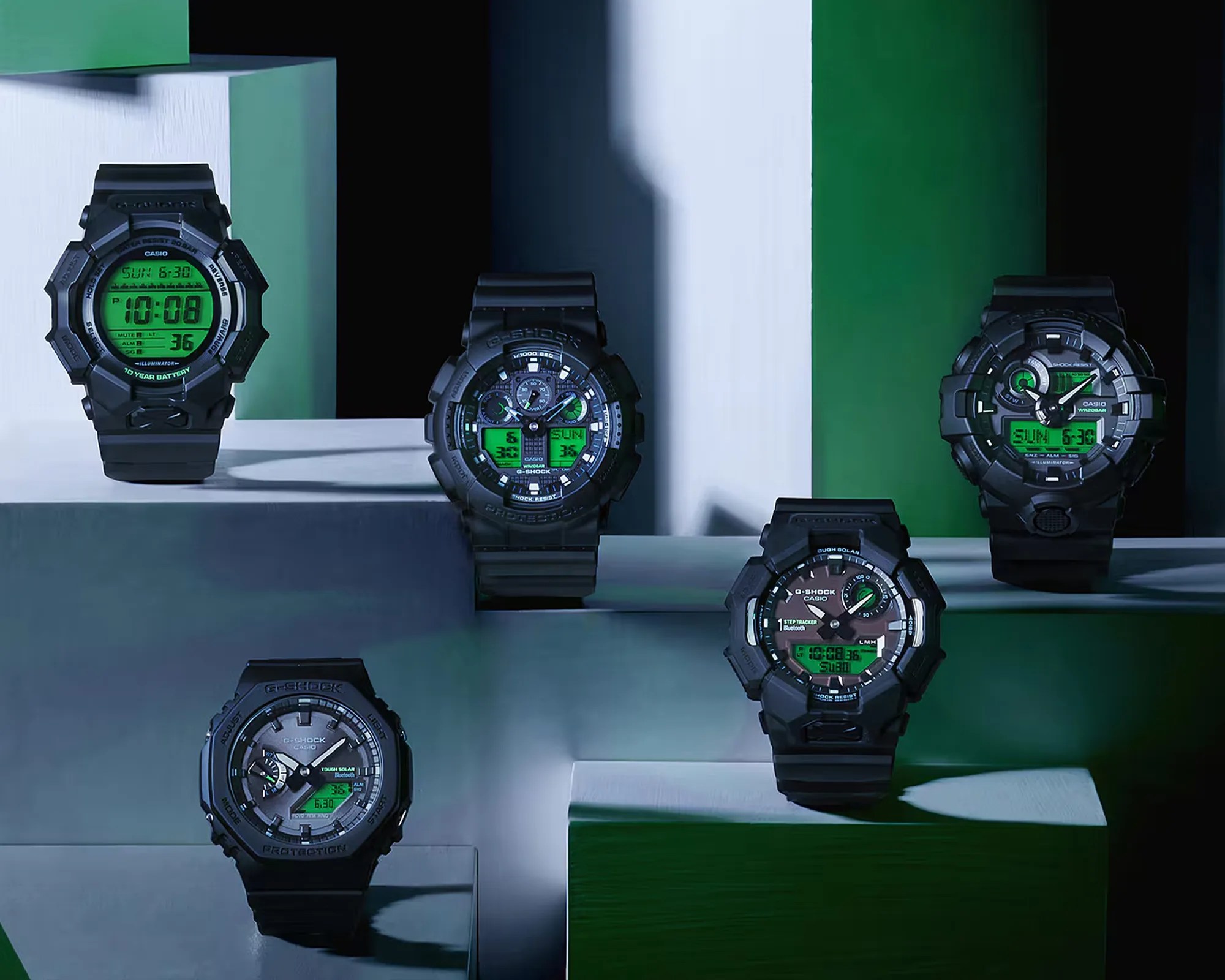 Five black Casio G-Shock digital-analog watches with green digital displays arranged on white and green geometric platforms.