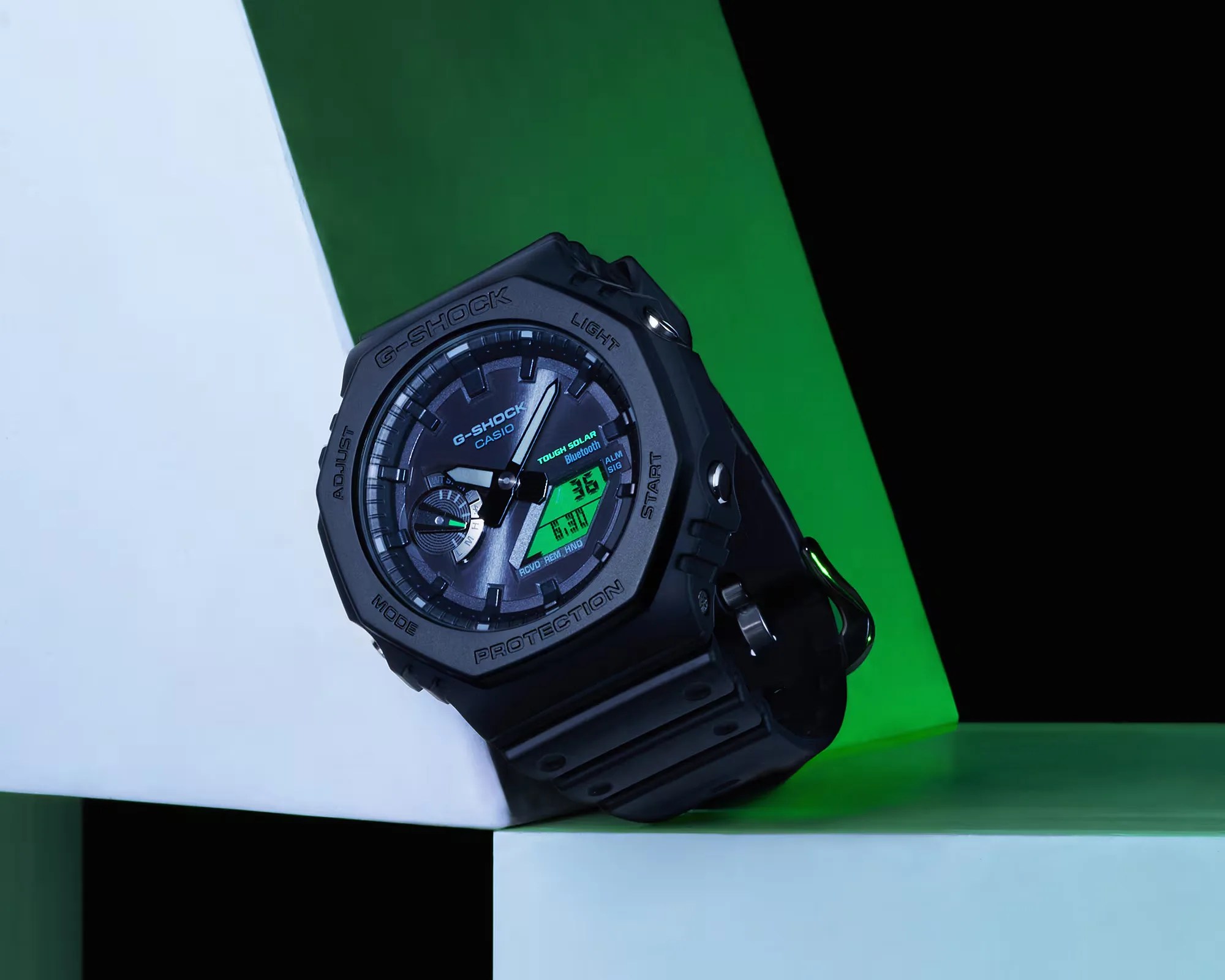 Black Casio G-Shock watch with analog-digital display and green backlight on geometric white and green blocks.