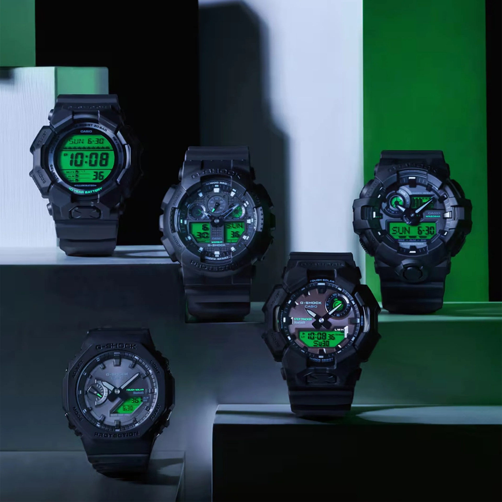 Five black Casio G-Shock digital-analog watches with green backlit displays arranged on dark and green geometric platforms.