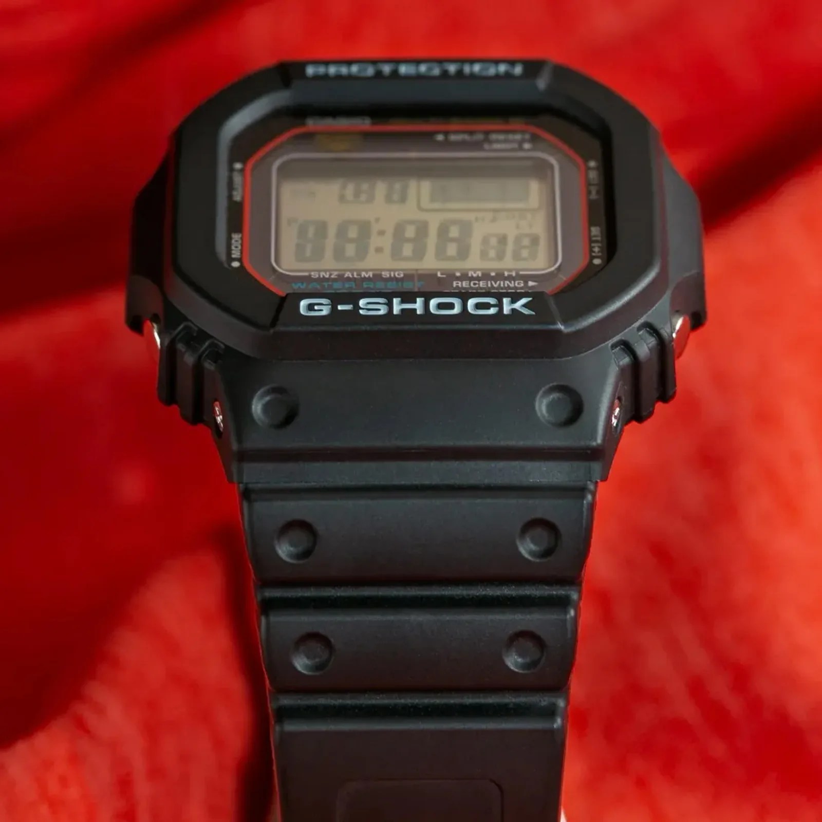 Black G-Shock digital watch with rectangular face and rubber strap on red fabric background.