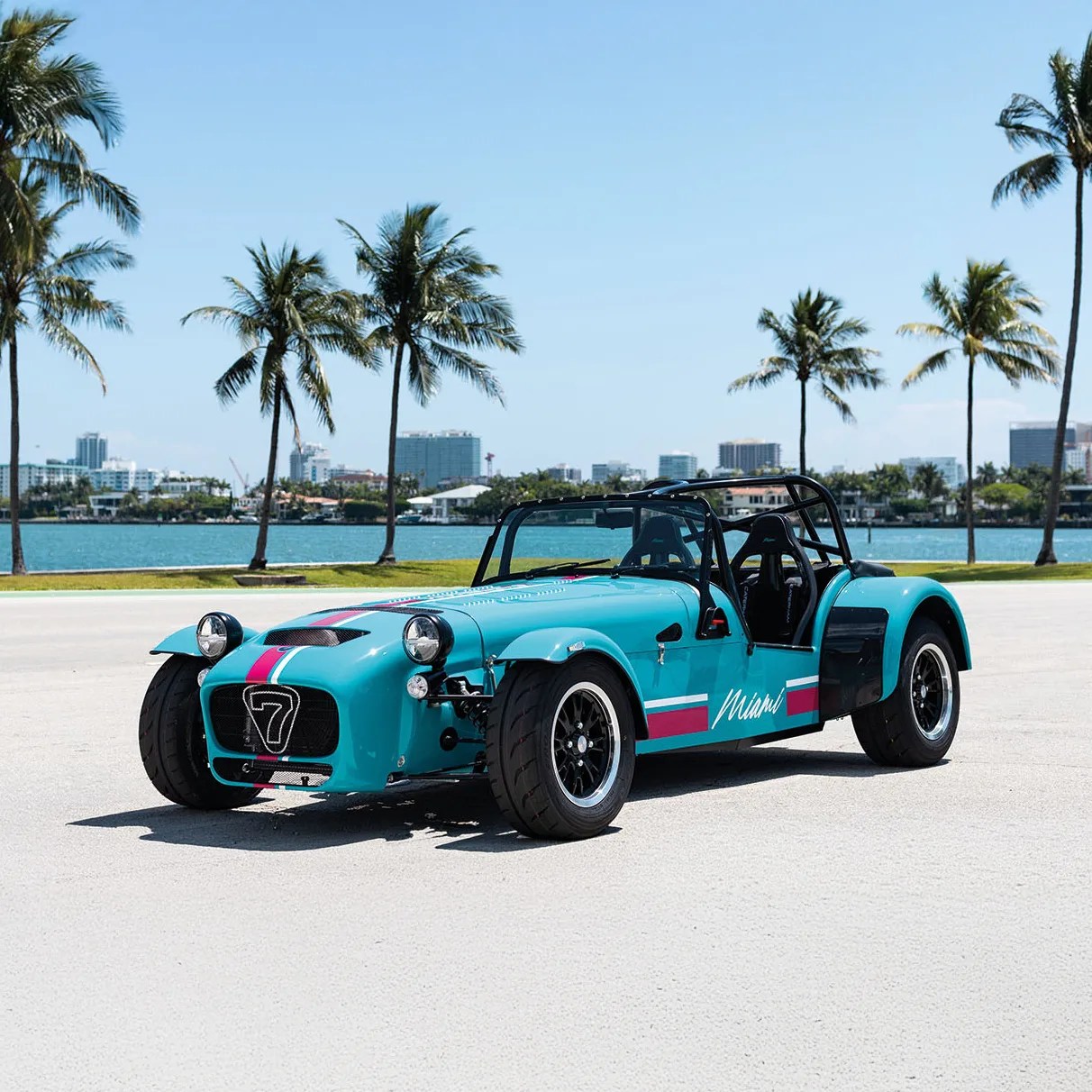Teal open-top sports car with black roll cage and "Miami" decal parked on a sunny waterfront road with palm trees.