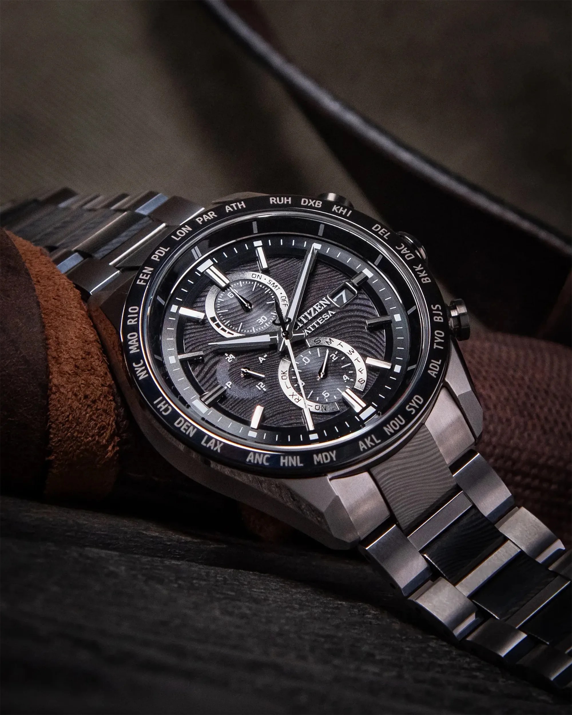 Black and silver Citizen wristwatch with multiple dials and city abbreviations on the bezel, on a brown fabric surface.
