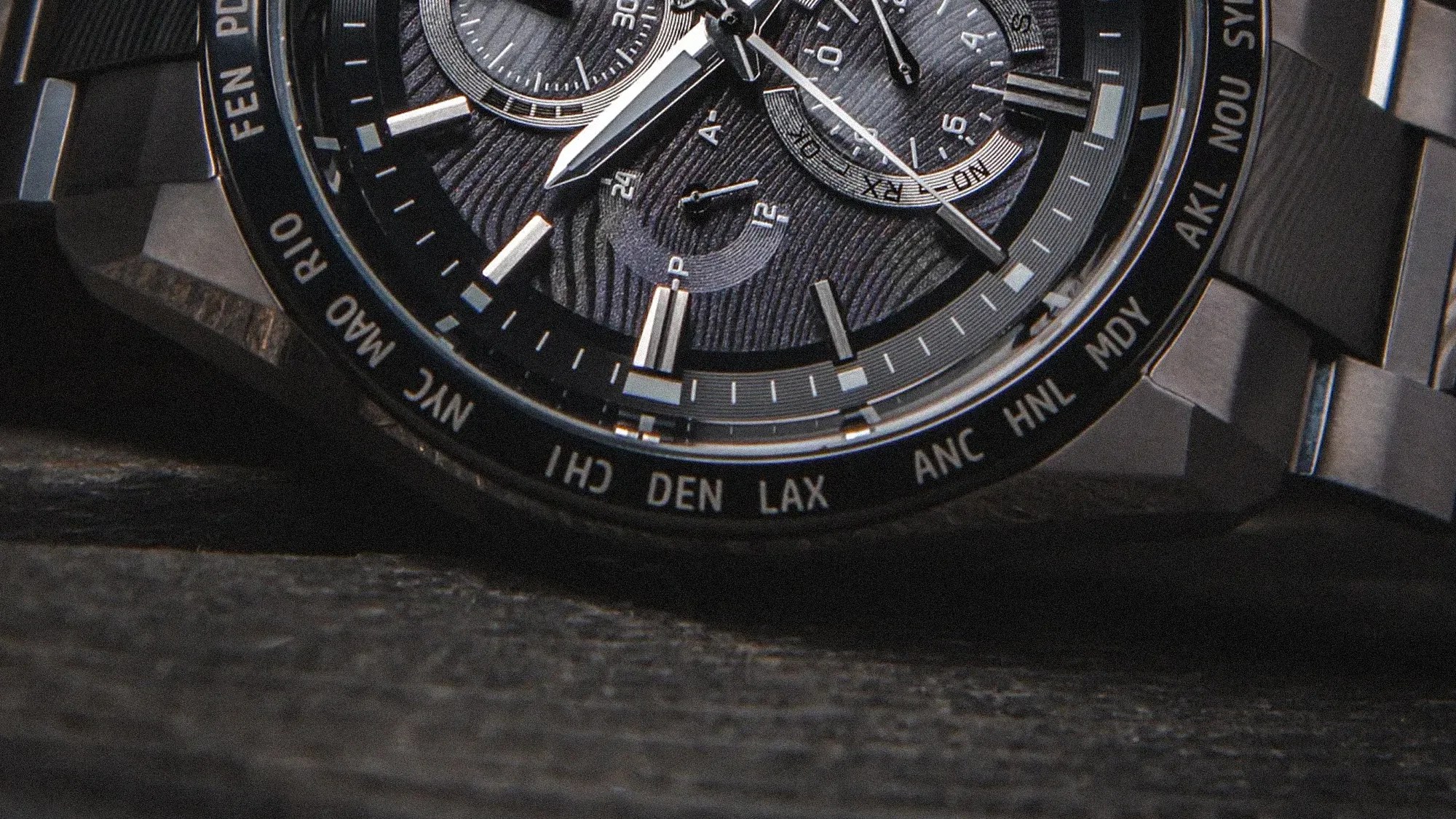 Close-up of a black watch face with silver hour markers and city abbreviations on the bezel.