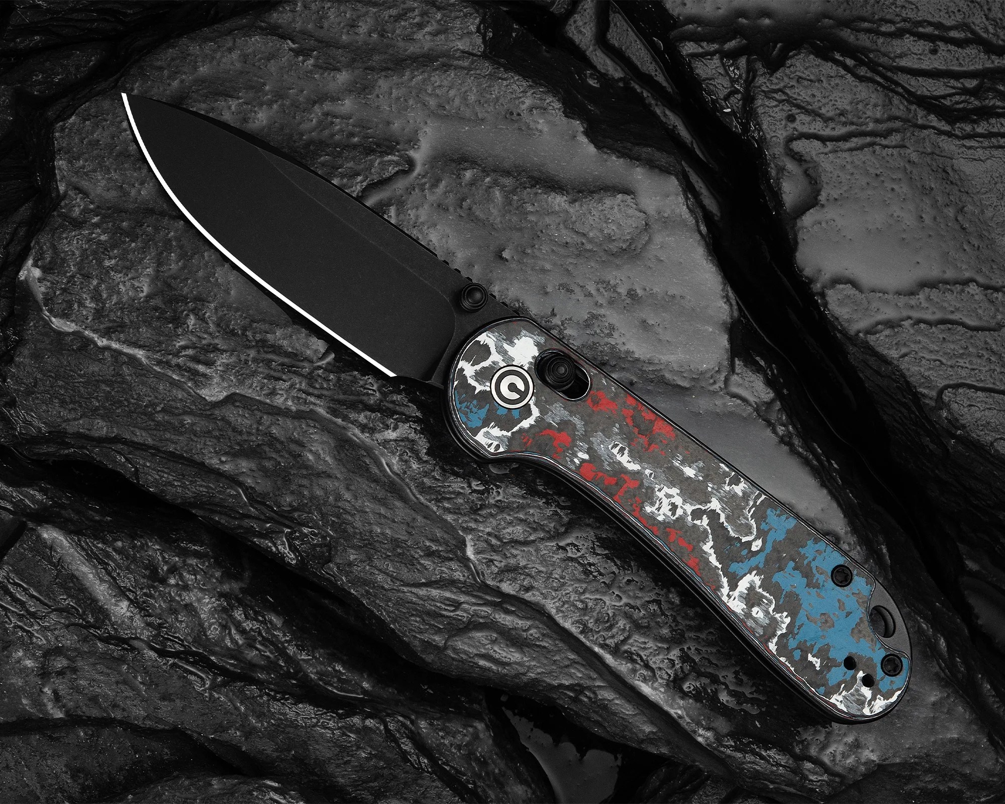 Folding knife with black blade and handle featuring red, white, and blue camouflage pattern on textured dark rock surface.