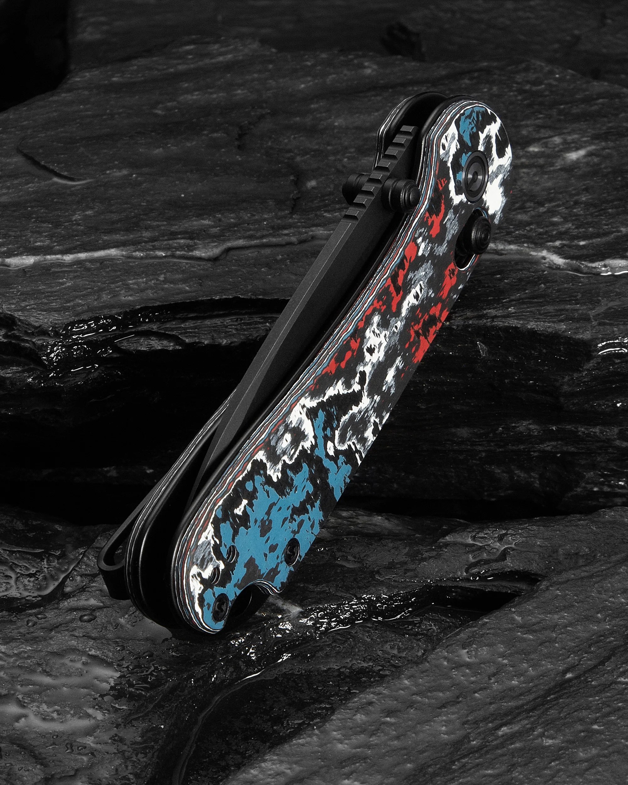 Folded pocket knife with a black blade and a handle featuring a red, white, and blue camouflage pattern on a dark textured surface.