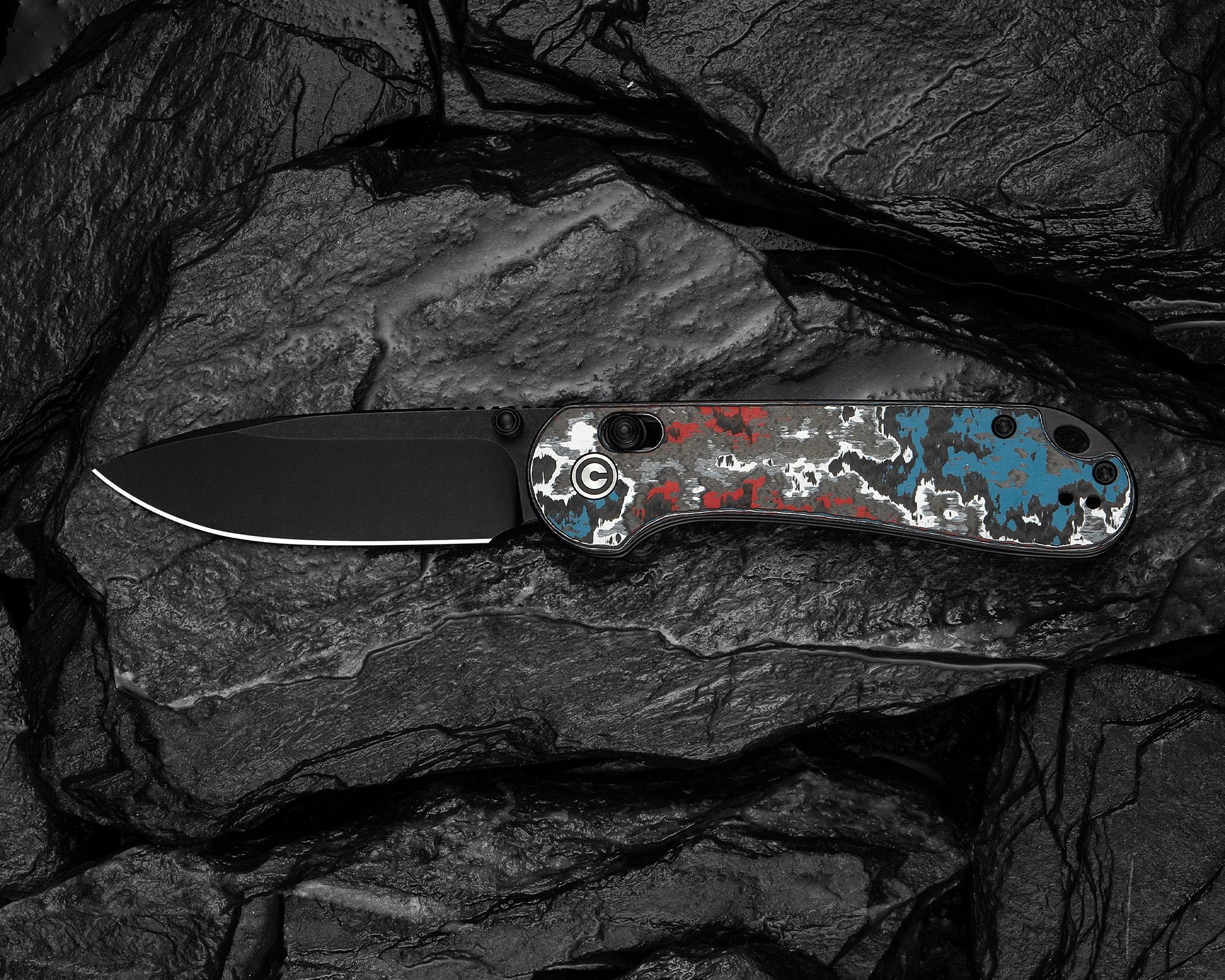 Black folding knife with a textured handle in red, white, and blue camouflage pattern on dark stone surface.