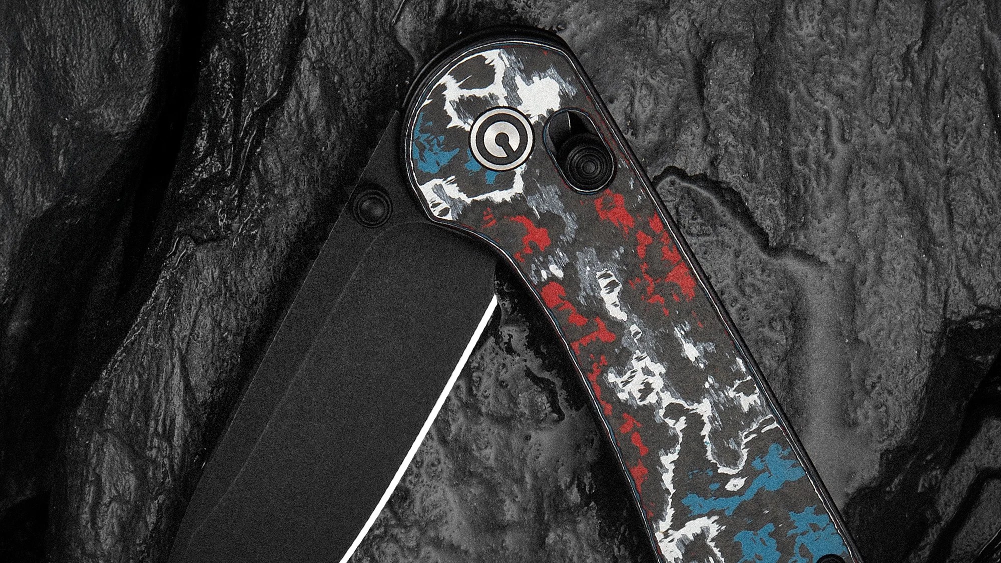 Folding knife with a black blade and a handle featuring a red, white, and blue camouflage pattern on a textured dark surface.