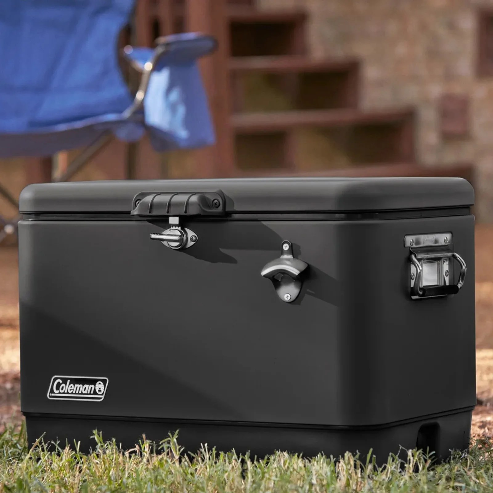 Black Coleman cooler with metal bottle opener and latch on grass outdoors.