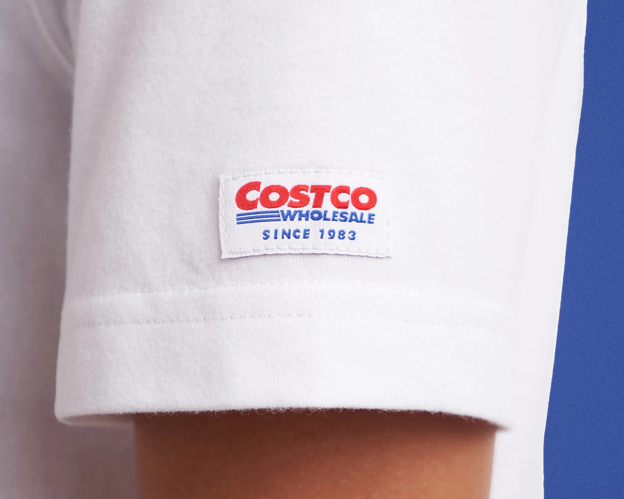 White fabric label with red and blue "Costco Wholesale Since 1983" text sewn on a white garment sleeve.