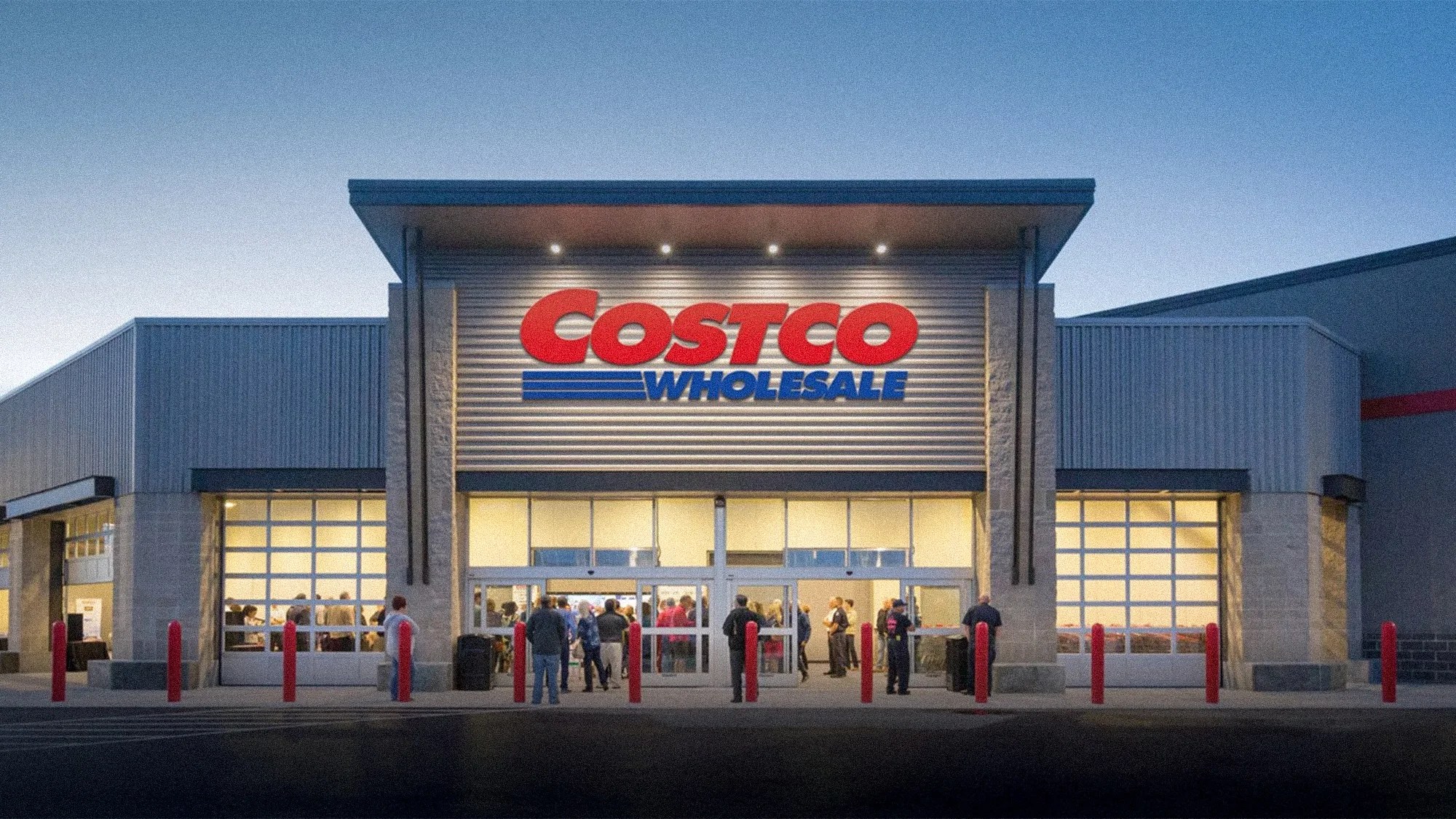 Costco Wholesale store entrance with illuminated sign and people gathered outside at dusk.