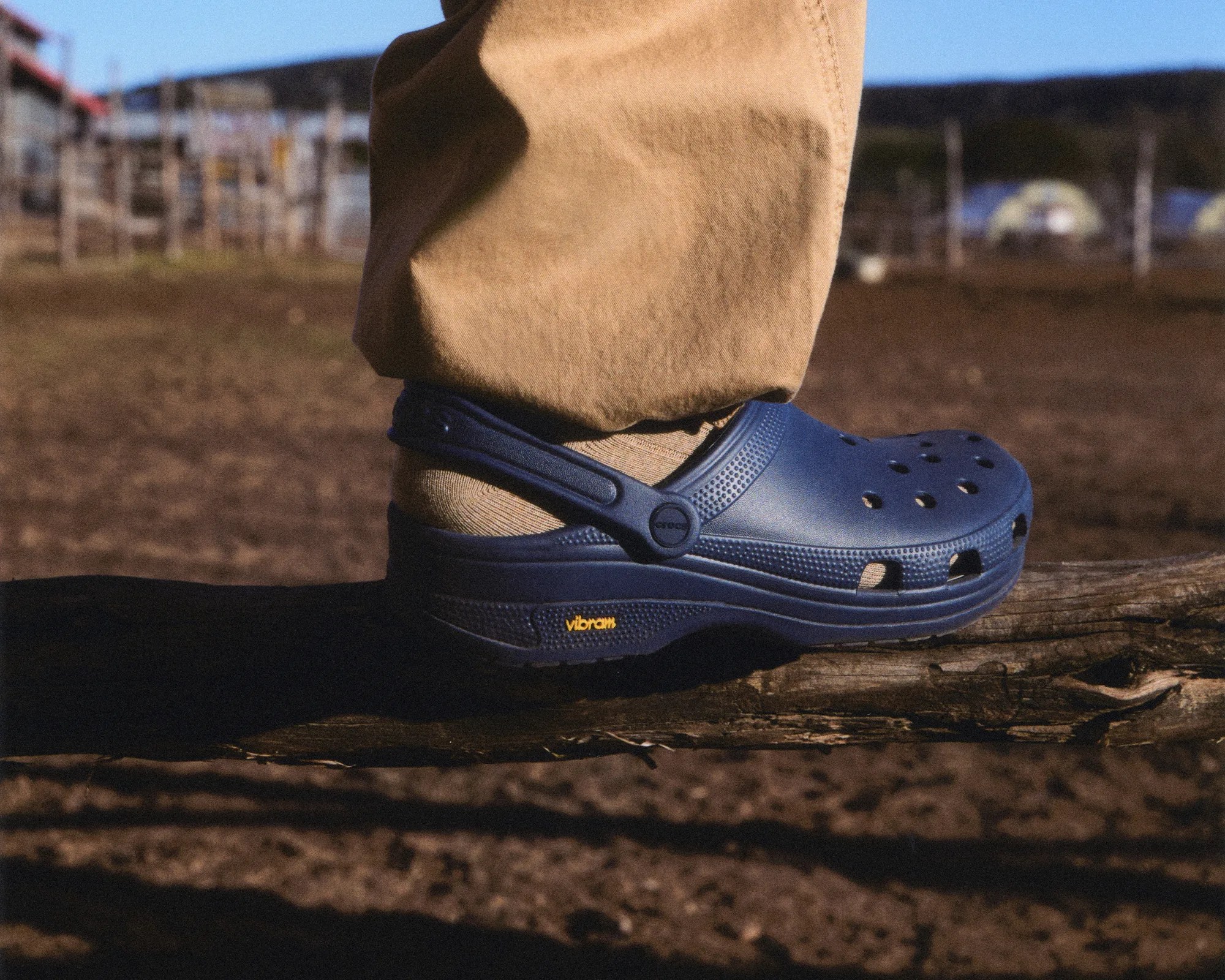 Navy blue Crocs with Vibram sole worn with beige socks and pants, standing on a wooden log outdoors.