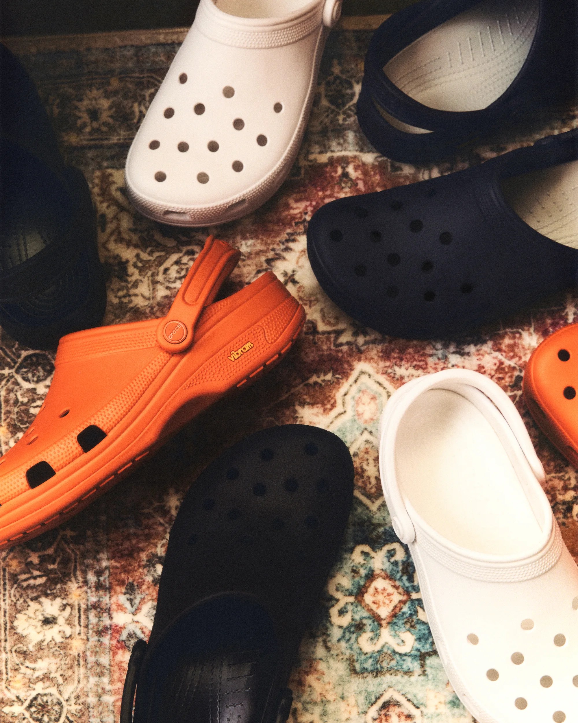 Various pairs of Crocs clogs in white, black, and orange arranged on a patterned rug.