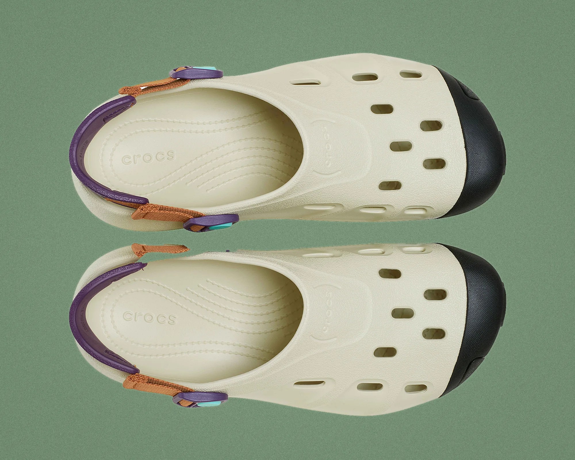 Pair of cream-colored Crocs with black toe caps, purple and brown adjustable heel straps on a green background.