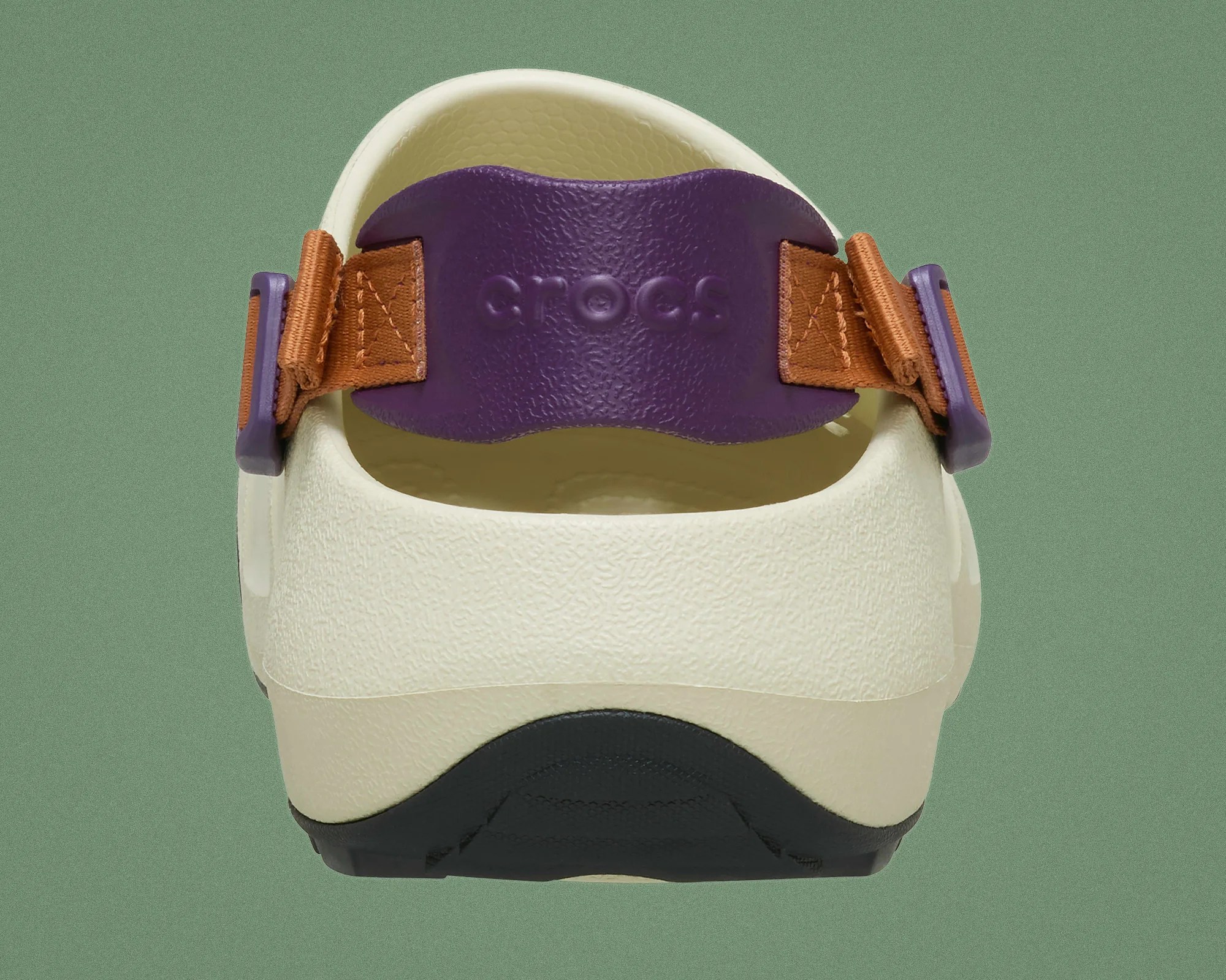 Back view of a cream Crocs shoe with a purple strap and brown adjustable fabric bands on a green background.