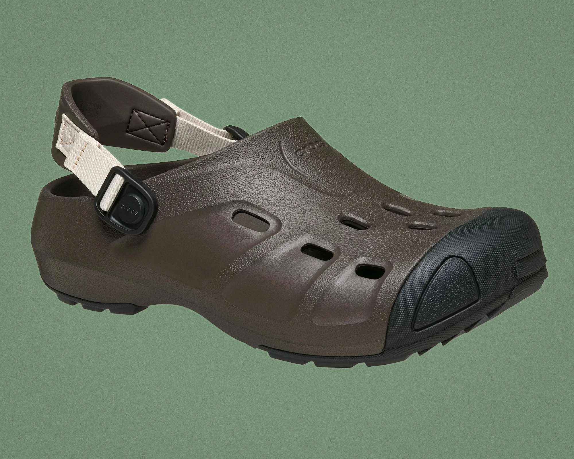 Brown Crocs clog with black toe cap and beige adjustable heel strap against a green background.