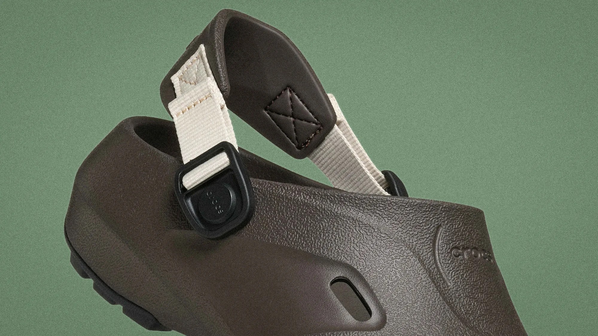 Close-up of a dark brown textured Crocs clog with an adjustable beige fabric strap and black buckle.