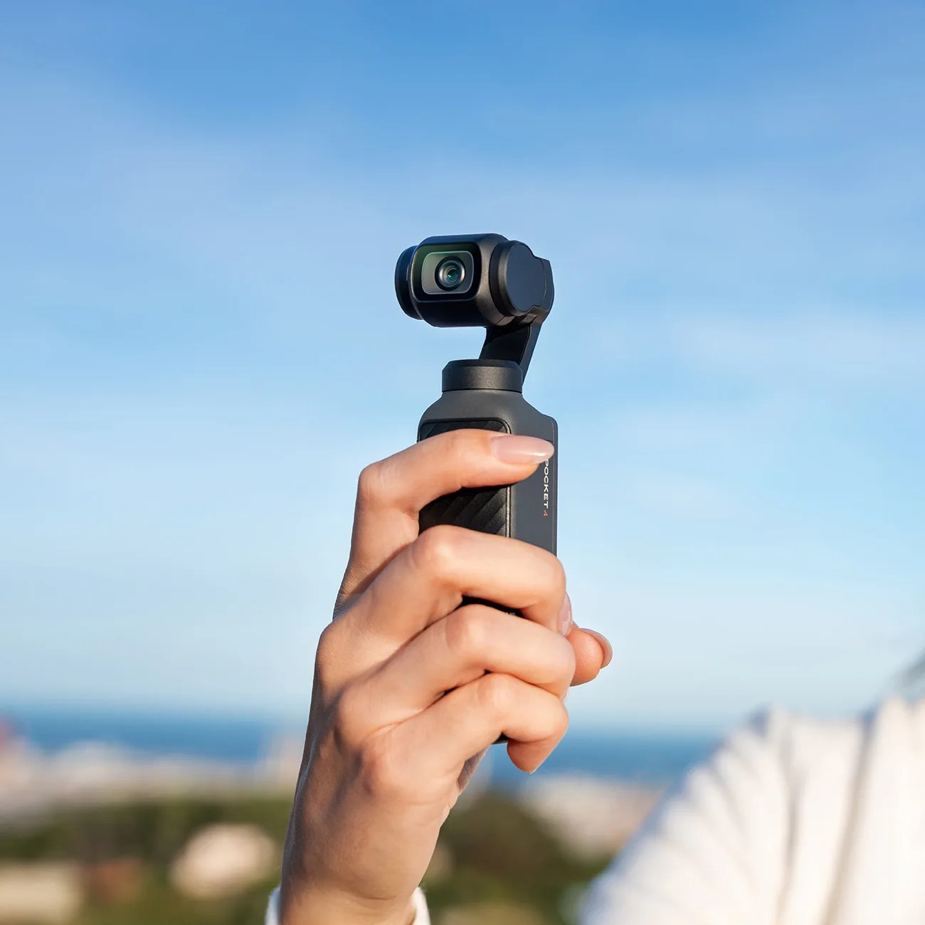 Hand holding a small black handheld gimbal camera against a blue sky background.