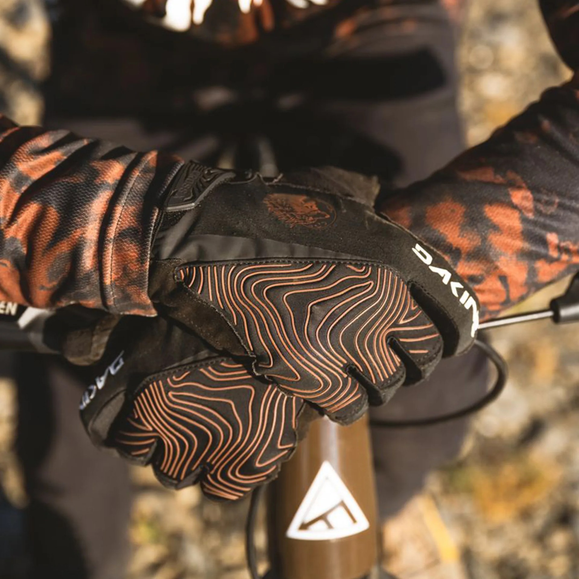 Black and orange Dakine gloves with topographic line patterns worn on hands gripping bicycle handlebars.